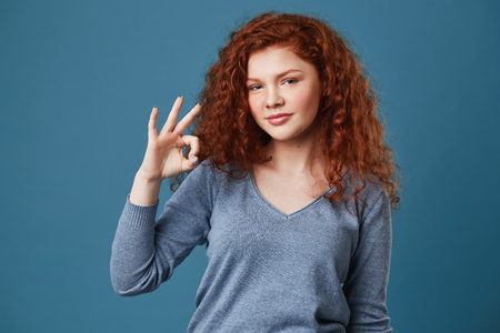 Cute young red haired girl with freckles in grey shirt showing OK gesture with hand, looking in camera with happy and relaxed expressionの写真素材
