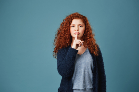 Portrait of good-looking ginger student girl with wavy hair holding index finger in front of lips showing friend not to tell story she saidの写真素材