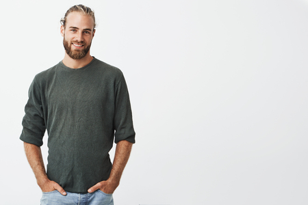 Handsome nordic man with beard and stylish hairstyle in grey shirt and jeans smiling, looking in camera, keeps hands in pockets.の写真素材