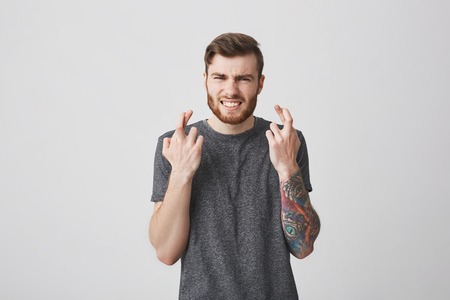 Waiting for competition results. Body language. Portrait of young attractive bearded tattooed male student with trendy short haircut crossing fingers, closing eyes with nervous face expression.の写真素材