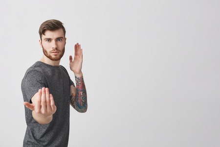Young good-looking serious bearded guy with stylish haircut and tattoo on left arm looking in camera with serious expression, making fight pose for funny party photo.の写真素材