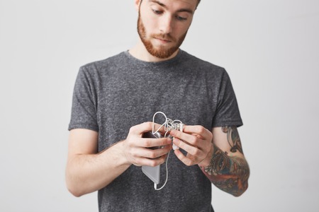 Close up of young good-looking unhappy caucasian bearded man with short dark hair and tattoo on arm in casual outfit holding in hands smartphone and earphonesの写真素材