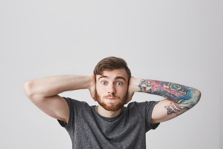 I dont hear anything. Attractive funny bearded man with fashionable hairstyle and tattooed arm closing ears with hands with popped eyes, looking in camera Body language.の写真素材