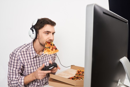 Portrait of young good-looking male gamer playing computer games with controller in headphones, holding pizza in mouth,looking in monitor with relaxed expression.の写真素材