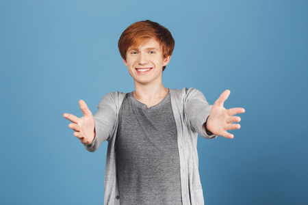 Come here, Ill give you a hug. Positive emotions. Redhead attractive joyful young guy in trendy grey clothes pulling hands in camera with bright smile and happy expression.の写真素材