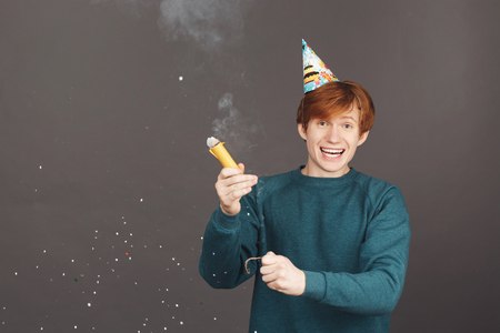 Positive emotions. Portrait of young red-haired guy in green sweater having fun on birthday party with family, blowing up flapper with happy and surprised expression. Black backgroundの写真素材
