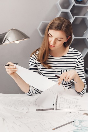 vertical portrait of young good-looking female designer with brown hair in striped shirt, looking at...
