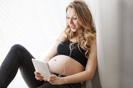 Close up of joyful young pregnant woman with blonde hair in black clothes sitting at window sill, talking with dad of future her baby on digital tablet in earphones. Happy family conceptの写真素材