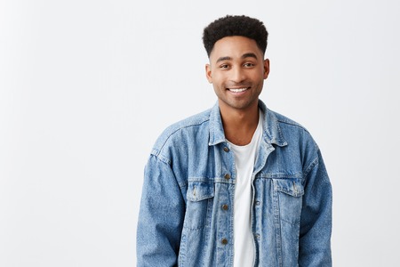 Close up isolated on white portrait of young beautiful cheerful dark-skinned male university student with afro hairstyle in white t-shirt and denim jacket smiling brightly in camera. Copy spaceの写真素材