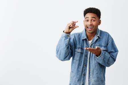 Little. Tiny. Isolated portrait of young good-looking funny happy black-skinned man with afro hairstyle in casual denim jacket gesticulating with hands, looking in camera with joyful expressionの写真素材
