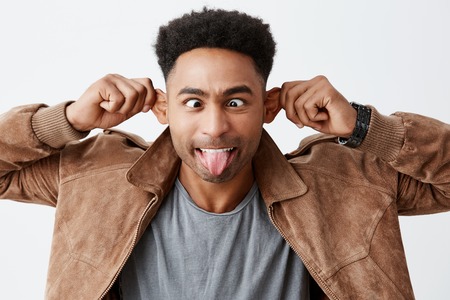 Playful mood Isolated portrait of mature dark skinned man with afro hairstyle in casual autumn clothes holding ears, mowing eyes, showing tongue in camera, tying make daughter laugh.の写真素材