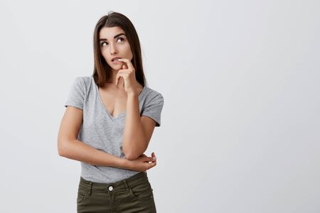 Close up of attractive sexy serious caucasian girl with dark long hair in casual gray shirt and jeans holding hand near mouth, looking aside with thoughtful and sneaky expression.の写真素材