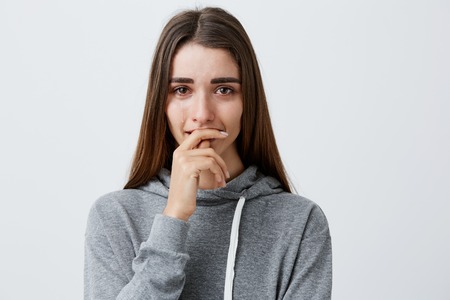 Negative emotions. Portrait of young charming pretty caucasian girl with brown long hair in fashionable casual hoodie crying, clothing mouth with hand, feeling depressed and crushed.の写真素材