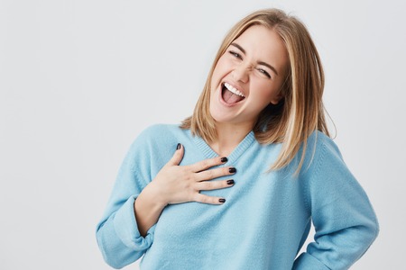 Happy beautiful girl with hair blonde hair smiling cheerfully with open mouth keeping hands on breast, pleased with unexpected gift from her boyfriend. Positive human expressions and emotionsの写真素材