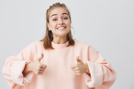 Excited blonde student girl showing thumbs up, showing her white teeth, demonstrating excellent results during exams at university. Young fair-haired woman looking at the camera in excitement.の写真素材