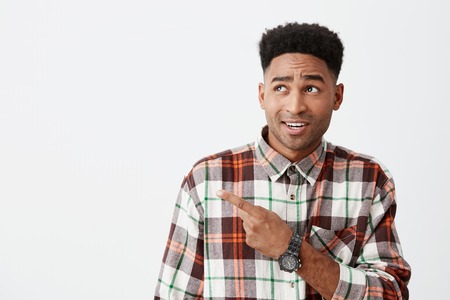 Close up of young handsome dark-skinned american male student with curly hairstyle in checkered long sleeve shirt pointing aside with index finger on white wall with cynical face expressionの写真素材
