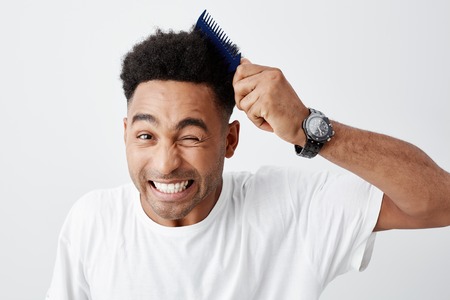 Curly hair problems. Close up of handsome young black-skinned american with afro haircut in casual white t shirt combing hair, looking in camera with funny face expression.の写真素材