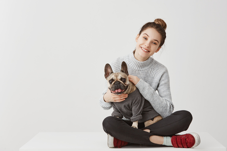 Portrait of happy young adult in good mood sitting on table at home crossed legs. Happy housewife fondling french bulldog with pleasure. Copy space, family conceptの写真素材