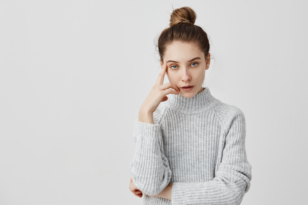 Young adult girl posing with annoyed look holding finger on temple. Attractive  woman in grey dress tired of listening uninteresting stories. Communication conceptの写真素材