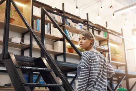Beautiful young blonde student girl with short hair in casual striped shirt spending time in modern library after university, preparing for exams with friends. Girl standing near stairs going to take some books,の写真素材