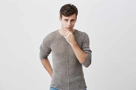 Pensive young caucasian guy wearing sweater touching his chin and looking ahead with faint smile....