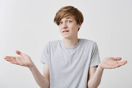 Caucasian young male with fair hair and blue eyes has hesitant and displeased expression, gestures doubtfully, being puzzled or confused. Uncertain european guy gestures against gray backgroundの写真素材