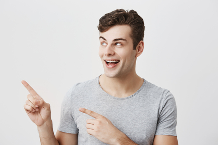 Portrait of caucasian handsome man having broad smile wearing gray t-shirt posing against studio wall pointing with index fingers at white copy space for advertisment or promotional content.の写真素材