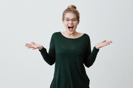 Amazed happy blonde girl dressed casually wearing eyeglasses exclaiming in excitement and shrugging shoulders in full disbelief after she received some unexpected postitive news. Body languageの写真素材