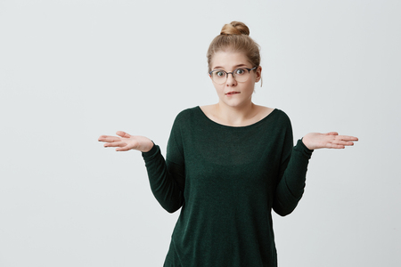 Clueless uncertain young woman with appealing appearance, blonde hair bun, shrugging shoulders, hesitates whether to go with friends on walk or study classes and preparing for exams. Life perceptionの写真素材