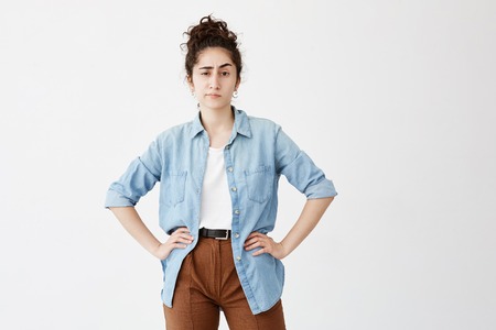 Young female employee resting at home after work. Pretty student girl with dark curly hair in bun, dressed casually waiting for her friends, standing with hands on hips against white backgroundの写真素材