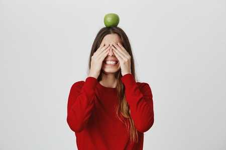 Healthy lifestyle. Positive emotions. Close up portrait of young beautiful cheerful caucasian girl with dark hair in red top clothing eyes with hands, holding apple on head, being excited.の写真素材