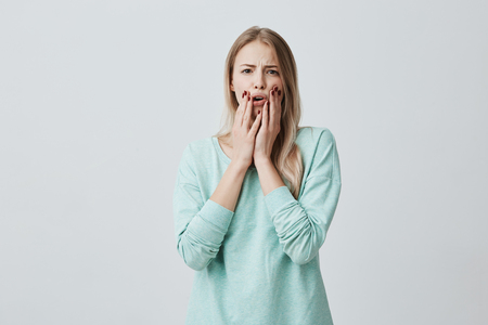 Shocked stunned blonde european woman with opened mouth, holding hands on cheeks, shocked by news she heard. Frightened terrified beautiful female model. Negative emotionsの写真素材