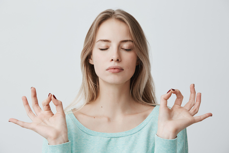 People, yoga and healthy lifestyle. Gorgeous young blonde woman dressed in light blue sweater keeping eyes closed while meditating indoors, practicing peace of mind, keeping fingers in mudra gestureの写真素材