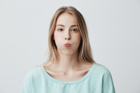 Sad blonde young woman has grumpy expression pouts her lips after quarrel with husband. Discontent dissatisfied female student looks gloomy after failing exam at university. Negative human emotions.の写真素材