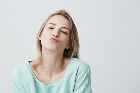 Young attractive caucasian woman dressed in blue casual clothes posing with kiss on lips with blonde dyed hair, having flirty look feeling confident and beautiful. Charming female having fun indoorsの写真素材