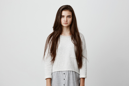 Brunette pretty charming young woman wearing loose sweater with dark straight long hair looking at camera with dark appealing eyes. Pretty girl posing against gray studio wall.の写真素材