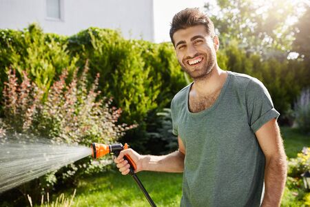 Close up outdoors portrait of young good-looking caucasian male gardener smiling in camera, watering plants, spending summer in countryside house.の写真素材