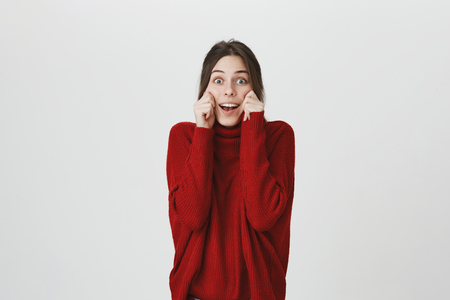 Happy cute modern girl in red trendy sweater holding her cheeks, expressing joy and happiness, isolated over white background. Forget about seriousness and remember to be childish sometimes.の写真素材