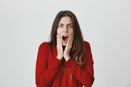 Shocked stunned emotional young woman in casual clothes with widely opened mouth, keeps hands on cheeks, is troubled and frustrated. European female can`t believe in shocking newsの写真素材