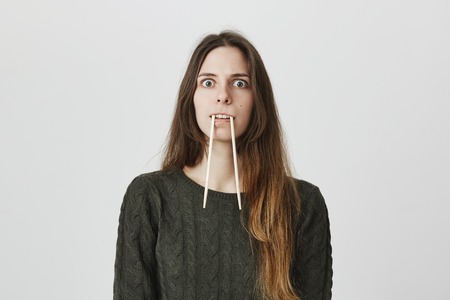 Portrait of funny childish adult female holding chopsticks in mouth imitating mammoth or beaver over white background. Girl plays guess game with friends trying to help them name animal she shows.の写真素材