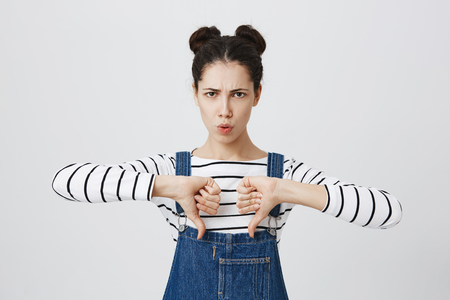 Portrait of brunette cute girl in denim overalls keeps thumbs down, disapproves something, pouts lips, frowns face in discontent. Pretty young female doesn`t like boring film.の写真素材