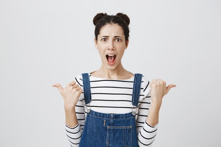 Amazed stunned female model with hairbuns, wearing denim overall, looking with puzzlement and...