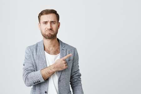 Dissatisfied male model with stubble, wearing gray jacket, looking at camera with sad expression, curving lips in displeasure, pointing with forefinger at copy space on studio wall.の写真素材