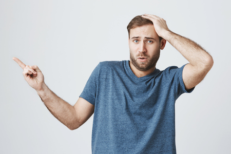 Portrait of scared amazed young man with stubble, looks with terrified expression at camera, indicates with forefinger at blank copy space on wall, isolated against gray concrete backgroundの写真素材