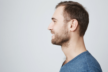 Close up profile of stylish smiling attractive male with stubble and dark hair wearing blue t-shirt posing against gray blank wall with copy space for your text or advertising contentの写真素材