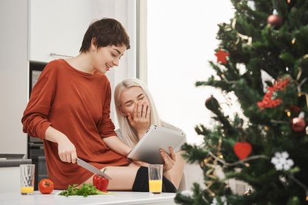 Side-view portrait of attractive skinny shirt-haired girl cutting vegetables and showing something in tablet to het friend who laughs out loud and enjoyes spending time with her.の写真素材