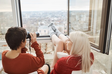 Back portrait of hot attractive women sitting on balcony with legs leaned on window, using binocular and drinking coffee. Girls fool around and spy after their neighbors or enjoy scenery of their townの写真素材