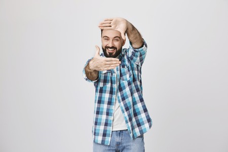 Studio shot of funny bearded european male office worker, holding hands in frame gesture while looking through it and smiling broadly, over gray background. Person acts like he is movie directorの写真素材