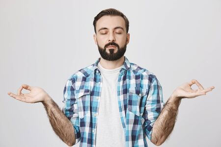 Studio shot of concentrated bearded adult man meditating in casual clothes, showing zen gesture with closed eyes, over gray background. In sound mind there is sound bodyの写真素材