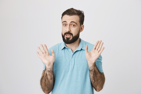 Picture of bearded man raising his eyebrows high, holding his palms up near white wall. Emotions and...